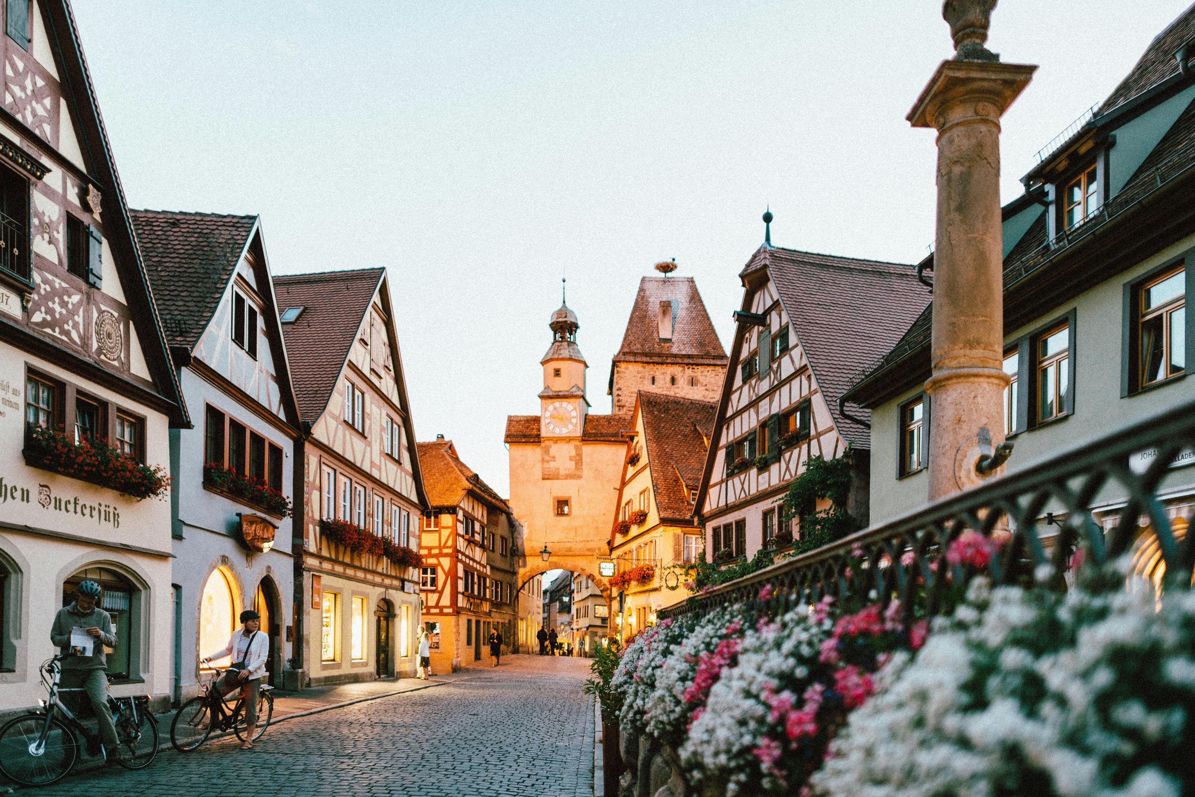 Charming European street in the evening with traditional half-timbered houses, flower-lined paths, and a historic clock tower in the background.